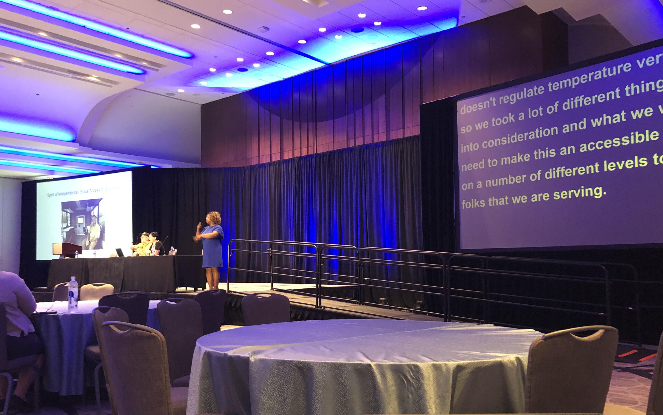 Picture of a hotel ballroom stage shown, with a large screen of PowerPoint on the left, a large screen of CART captions on the right, and an ASL interpreter standing in the middle of the stage.